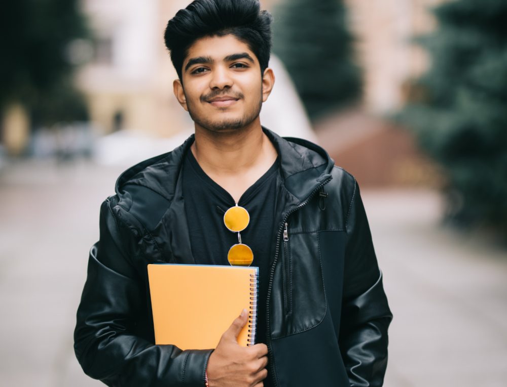 handsome-young-indian-student-man-holding-notebooks-while-standing-street